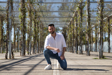 Handsome young man with beard, wearing white shirt and jeans, with one knee on the ground and hands on legs. Concept fashion, beauty, modern, trend, model.