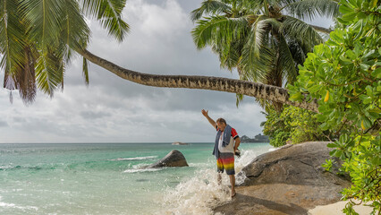A man stands on a boulder on the beach, smiling. The hand is raised. Turquoise waves of the ocean break on a stone. Foam and splashes all around.  A palm tree leaning over the water, against a sky.