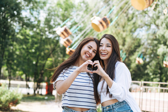 Young women with long hair friends having fun showing heart with theit hands at amusement park