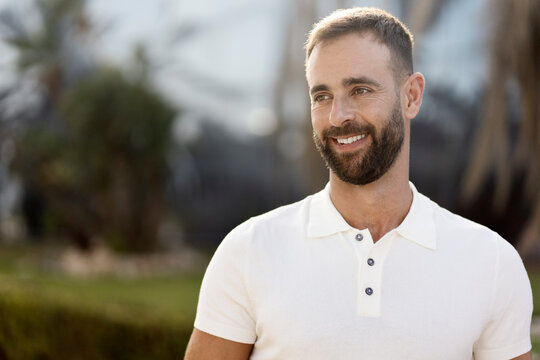 Confident smiling hispanic man wearing white stylish t shirt  looking away standing on the street, copy space. Portrait of middle aged bearded fashion model posing for pictures, sunny day