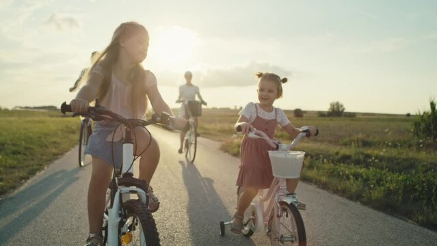 Caucasian family of two elementary age girls on foreground and parents in the background riding bikes on village road. Shot with RED helium camera in 8K.  