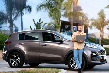 Pensive middle aged latin man using laptop computer, working online standing near new car. Handsome...
