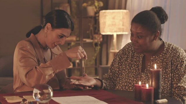 Waist up slowmo of fortune teller using pendulum over hand of African American woman came to spiritual seance