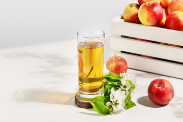 Apple juice. Full glass of natural apple juice on a wooden podium with red apples and blossom branche on a white table with copy space. Summer natural drink. Soft focus stile