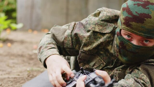 A young soldier in a mask aims for shooting from a machine gun while sitting in ambush, close-up