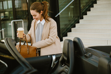 Young woman smiling and drinking coffee while standing by her car