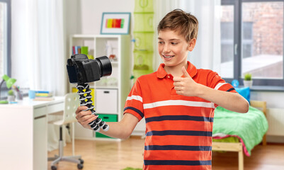 blogging, videoblog and people concept - portrait of happy smiling boy video blogger with camera and tripod videoblogging and showing thumbs up over home room background
