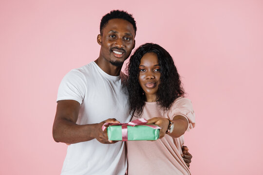 Cheerful beautiful african american couple showing their gift over pink studio background. African lovers exchanging gifts while anniversary celebration. - Powered by Adobe