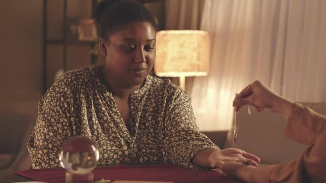 Waist up slowmo of fortune teller using pendulum while studying palm of young Black woman during seance