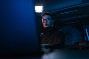 cybercrime, hacking and technology concept - young man in glasses writing code on laptop in dark...