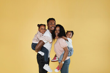 Time with family. Overjoyed african american family laughing and posing isolated over yellow studio wall. Cheerful father and mom carrying their daughters on their back.