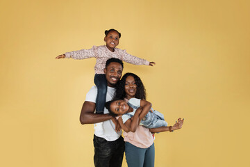 Cheerful african man holding excited daughter on hands. Woman holding another daughter looking posing and looking at camera, standing isolated over yellow studio background.