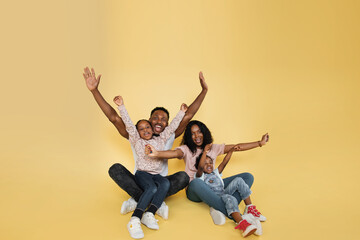 Family fun. Young african american parents spending time with their little daughters with open arms, laughing together over yellow background, banner, panorama, copy space.
