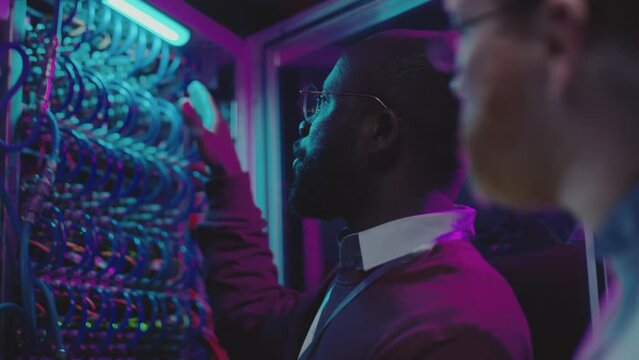 African American IT engineer checking server cables and talking to Caucasian male colleague as he working on digital tablet in data center