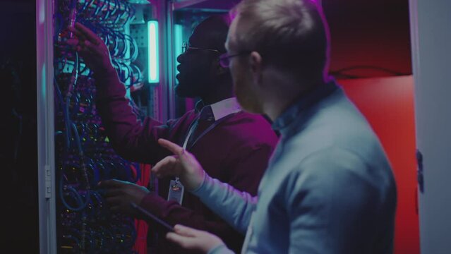 African American network technician checking server cables and speaking with Caucasian male colleague as he using digital tablet in data center