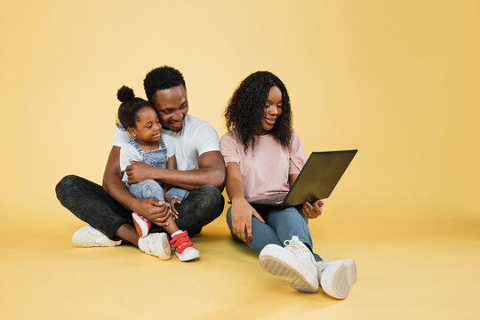 Happy family, technology, communication concept. Young african parents sitting on floor with their daughter and embracing, using laptop for video call, over yellow studio background, free space.