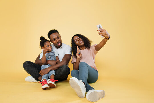 Portrait of smiling african american family of three sitting on floor with smartphone while taking funny picture of them showing tongues over yellow studio background, free copy space, banner.