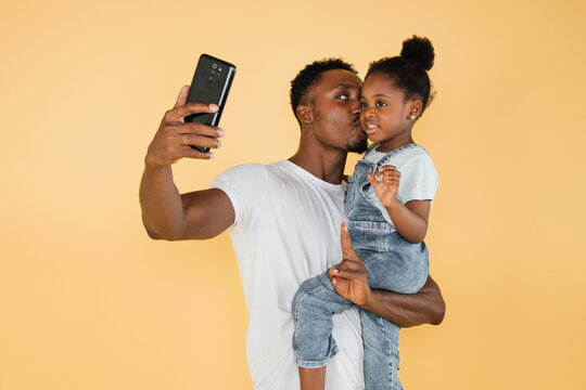 Cheerful african american dad kissing his daughter girl in casual clothes isolated on yellow background studio portrait. People lifestyle concept. Mock up copy space. Doing selfie shot on mobile phone