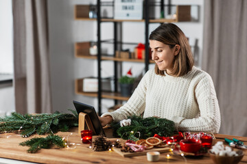 winter holidays, diy and hobby concept - happy smiling woman with tablet pc computer making christmas fir wreath at home