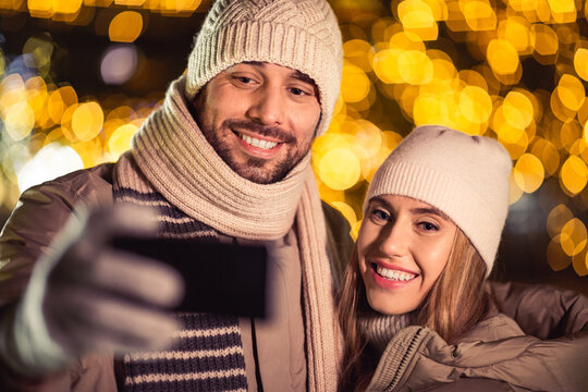 winter holidays and people concept - happy smiling couple taking selfie with smartphone and hugging over christmas lights in evening