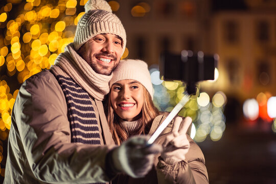 winter holidays and people concept - happy smiling couple taking picture with smartphone on selfie stick and hugging over christmas lights in evening