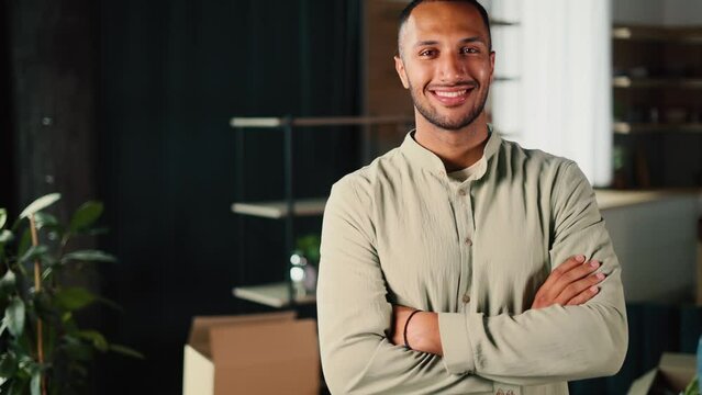 Portrait of attractive African american man standing in living room after moving in with his wife who unpacking boxes in the background. New home.