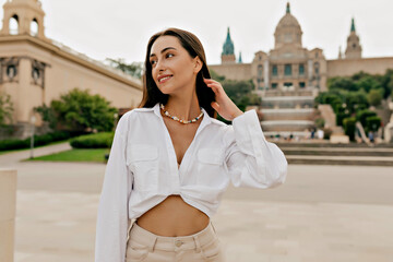 Adorable good-looking slim woman with tanned skin and long hair wearing white blouse looking away and walking around in the city in warm sunny day