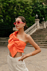 Good-looking european female model in pink fashion glasses wearing orange stylish blouse and beige jeans looking straight and walking outdoor in warm summer day