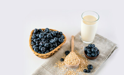 Fototapeta na wymiar Raw oatmeal flakes with black blueberries and a glass of milk. High angle of fresh blueberries and a wooden spoon with oatmeal placed on table near glass of milk and on white background in light room