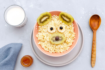 Funny cute kids childrens baby's healthy breakfast lunch oatmeal porridge in bowl look like bear,mouse face decorated with kiwi, banana, dried berry fruits,milk,honey. dessert food art on gray table