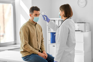 Obraz premium medicine, healthcare and people concept - female doctor in mask with infrared forehead thermometer measuring temperature of man patient at hospital