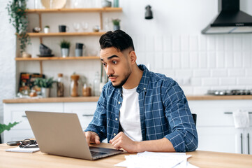 Surprised puzzled mixed race young man, freelancer or creative manager, sitting at home in the kitchen, shocking looking at the laptop screen, in stress of unexpected news, opened his eyes wide