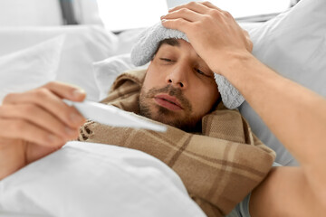 people, health and fever concept - sick man with cold compress on his forehead measuring temperature by thermometer lying in bed at home
