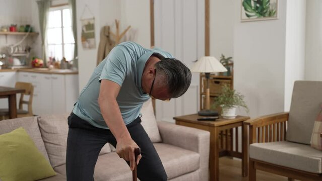 asian injured mature male groaning in pain while he tries to get up with a stick after fall at home. He holds his lower back and can barely stand up