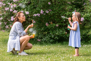 Obraz premium family, motherhood and people concept - happy mother with little daughter blowing soap bubbles at summer park or garden