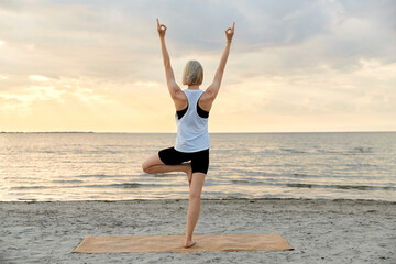Fototapeta na wymiar fitness, sport, and healthy lifestyle concept - woman doing yoga tree pose on beach over sunset