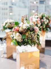 An exotic bouquet of protea, dry hydrangea, white and cream roses, barberry twigs, and leaves stands in a glass vase on a golden stand. Panorama of the city, floral decor and white candles in the back