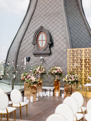 The decor of the outdoor wedding is on the roof against the backdrop of a classic brown tower with an oval window: gold decor, white chairs, bouquets of flowers, candles on the floor, and garlands.