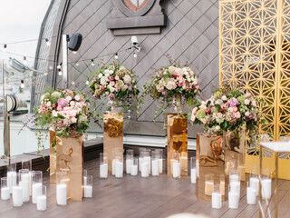 The decor of an outdoor wedding is on the roof against the backdrop of a brown tower with a window. Bouquets of white and pink flowers stand on gold stands, white candles in candlesticks on the floor.