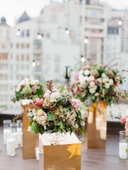 An exotic bouquet of protea, dry hydrangea, white and cream roses, barberry twigs, and leaves stands in a glass vase on a golden stand. Panorama of the city, floral decor and white candles in the back