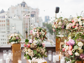 An exotic bouquet of protea, dry hydrangea, white and cream roses, barberry twigs, and leaves stands in a glass vase on a golden stand. Panorama of the city, floral decor and white candles in the back