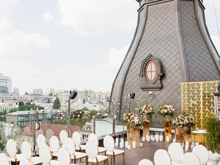 The decor of the outdoor wedding is on the roof against the backdrop of a classic brown tower with an oval window. Gold decor, white chairs, bouquets of flowers, candles on the floor, garlands.