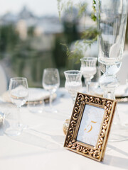 Number 5 of the banquet table in a golden frame stands on a banquet table on a white tablecloth. In the background are wine glasses, plates, and a tall glass vase.