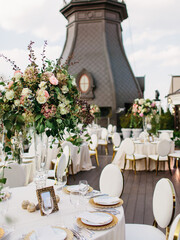 The decor of the wedding banquet is white on the roof against the backdrop of a brown tower with an oval window. On the table is a white tablecloth, cutlery, plates, wine glasses, bouquets of flowers.