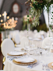 Wedding table setting in gold and white. On the table is a white tablecloth, dishes, cutlery, and a bouquet of flowers in a tall glass vase. In the background is a brown tower with an oval window.