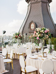 The decor of the wedding banquet is white on the roof against the backdrop of a brown tower with an oval window. On the table is a white tablecloth, cutlery, plates, wine glasses, bouquets of flowers.