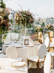 Served banquet tables for a wedding dinner on the rooftop. Table decor in white tones, a bouquet of roses, hydrangeas in a tall glass vase. In the background is a panorama of the city.