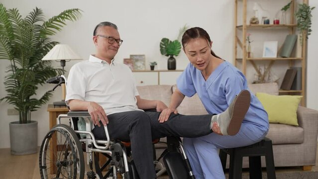 Chinese female nursing aide assisting elderly male on wheelchair to exercise his leg by raising it up gently. in-home care and physiotherapy for disabled senior concept