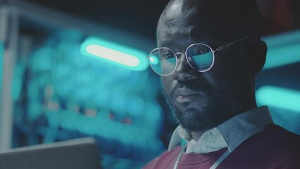 Tilt up shot of black IT professional working on laptop in dark server room of data center