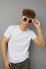 a man stands in a casual T-shirt on an isolated gray background with sunglasses on and holding them with his hand looks at the camera. Vertical Studio Photography